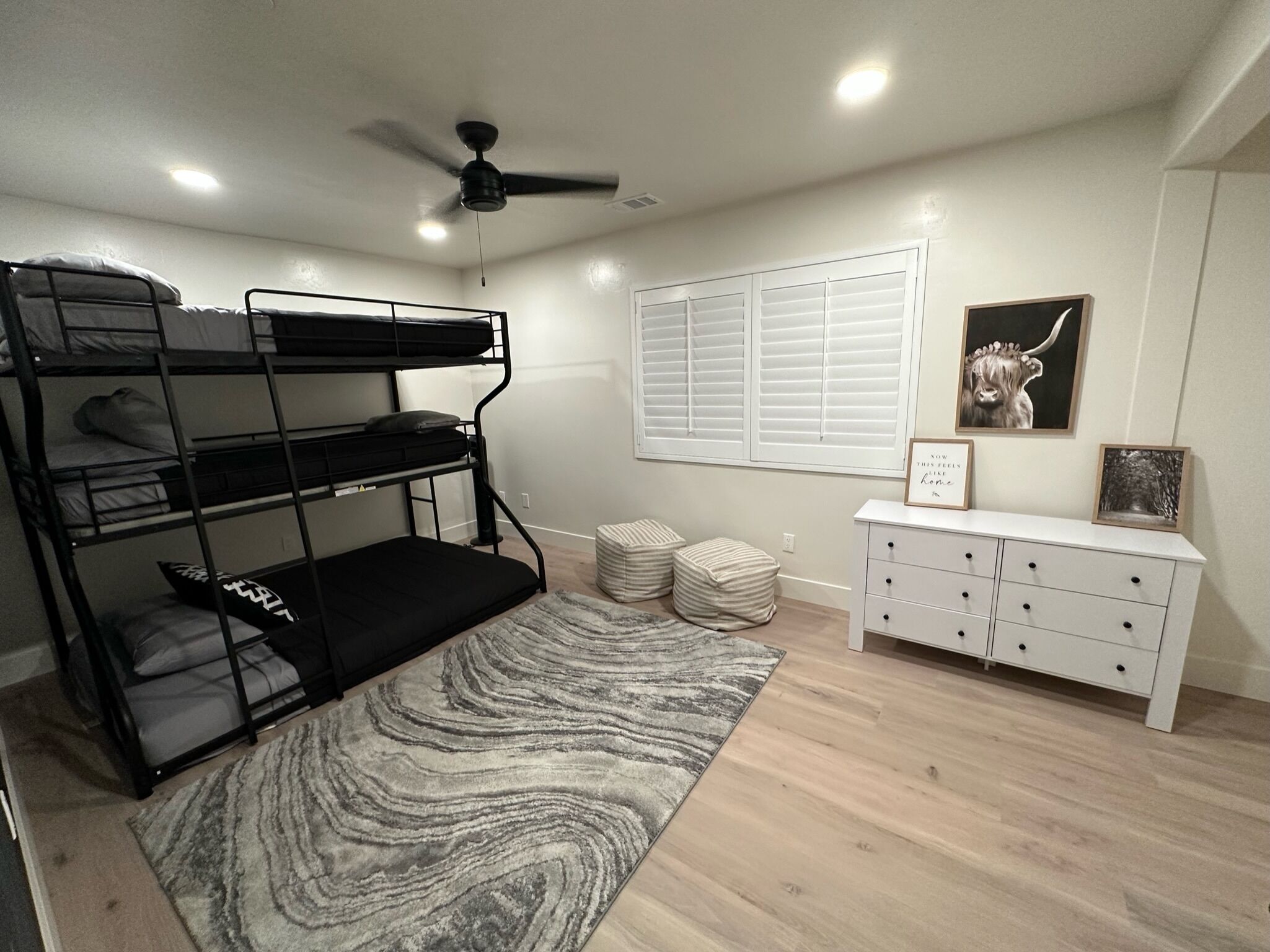 A bunk bed room with a ceiling fan, a window with blinds, a white dresser, and a framed picture on the wall.