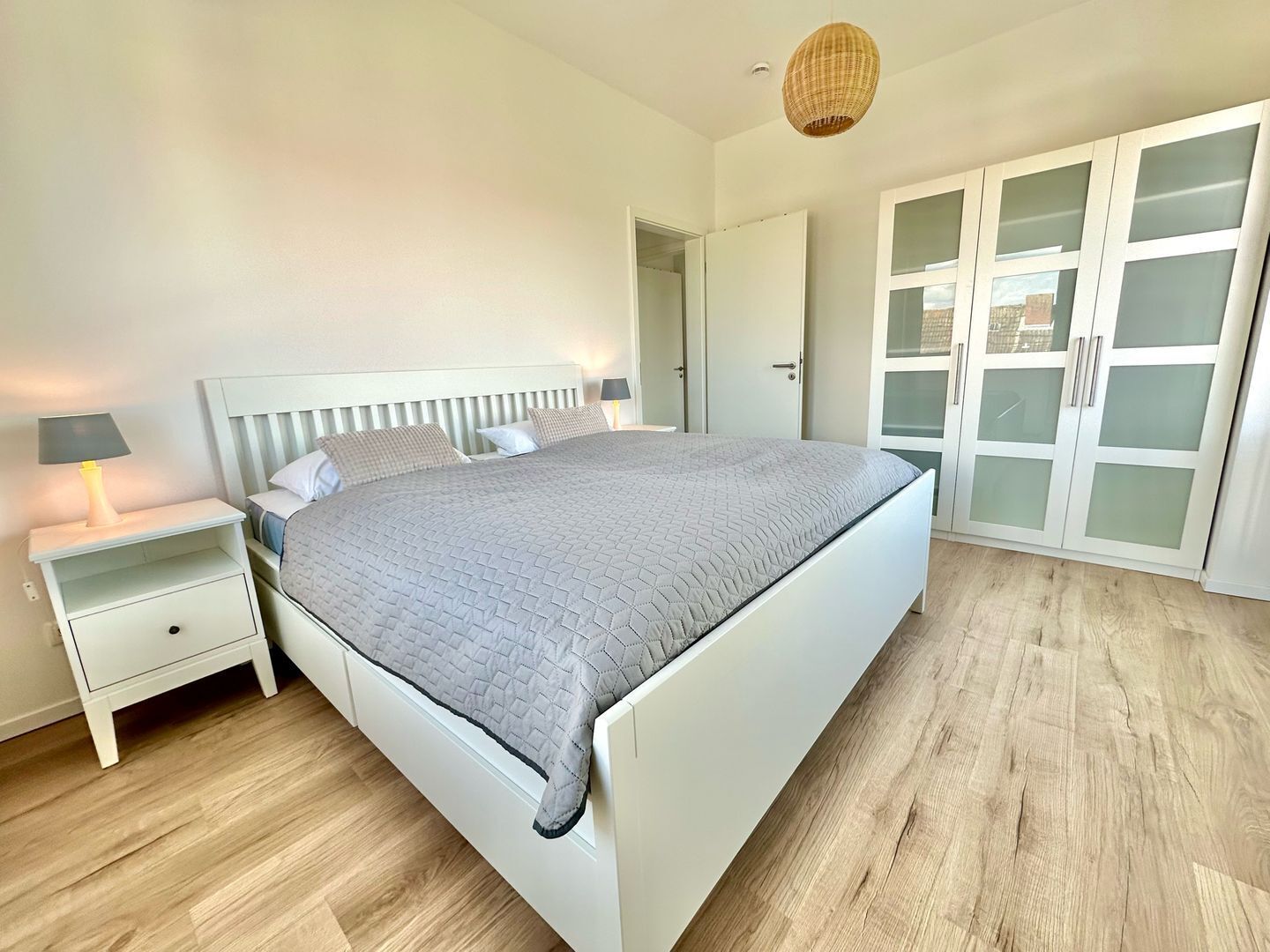 Un dormitorio con cama, mesitas de noche, lámpara, armario con puertas de cristal satinado y suelo de madera.