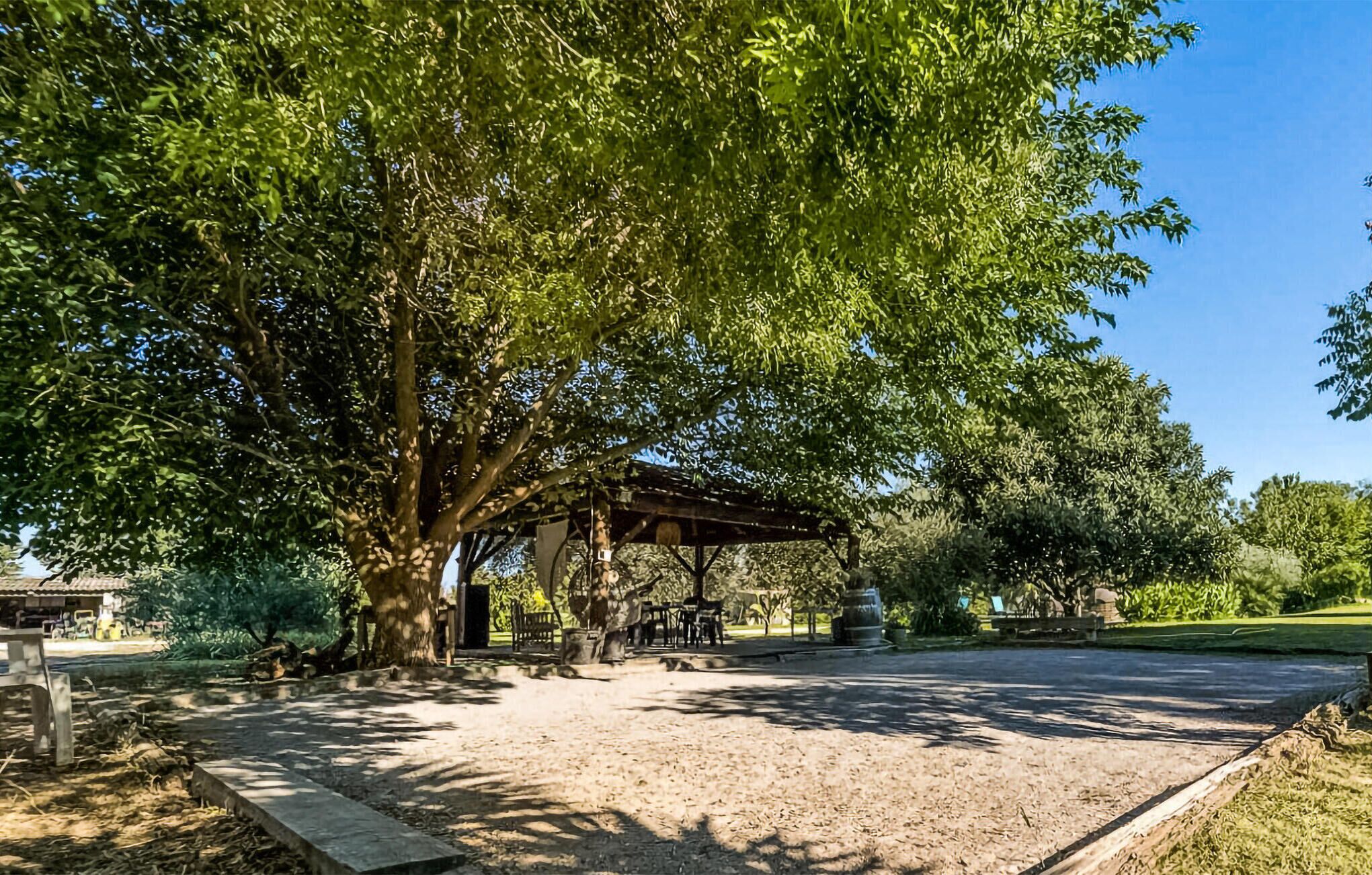 Un espace extérieur aménagé avec des tables et des chaises, protégé par un toit, et entouré d’arbres, avec un ciel dégagé au-dessus.