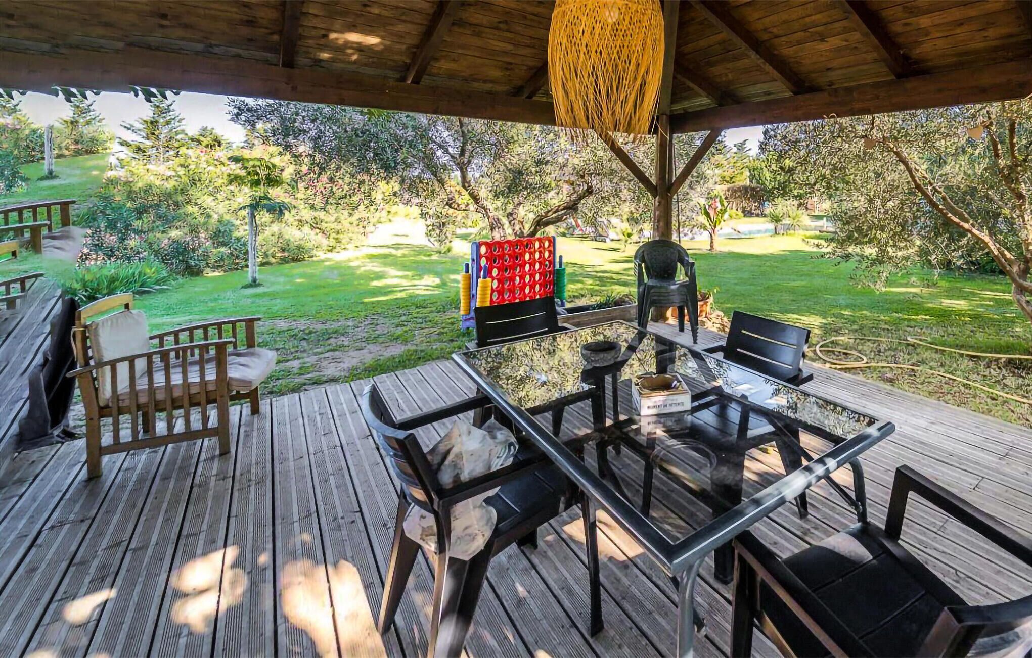 A wooden deck with a glass table and chairs, surrounded by greenery and trees.