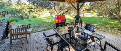 A wooden deck with a glass table and chairs, surrounded by greenery and trees.