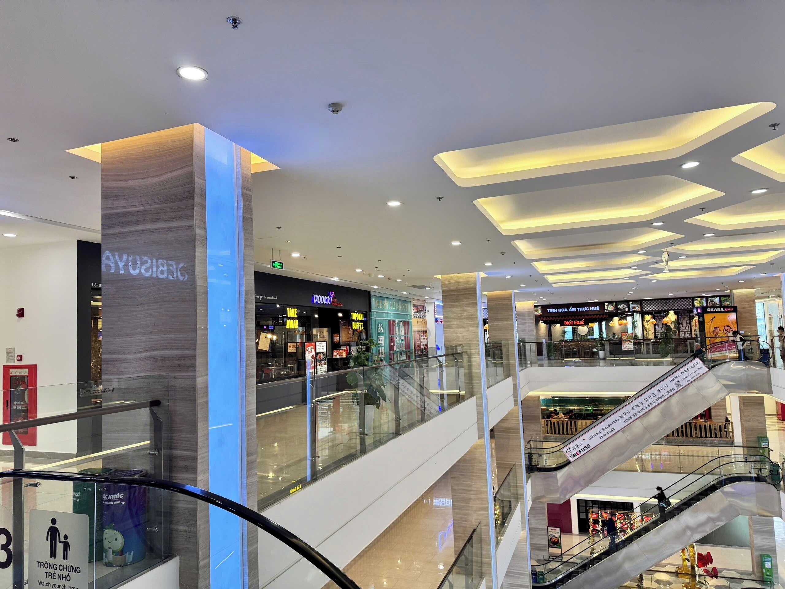 Un centro comercial de varios niveles con barandillas de vidrio, escaleras mecánicas y diversas tiendas, incluyendo una cafetería.