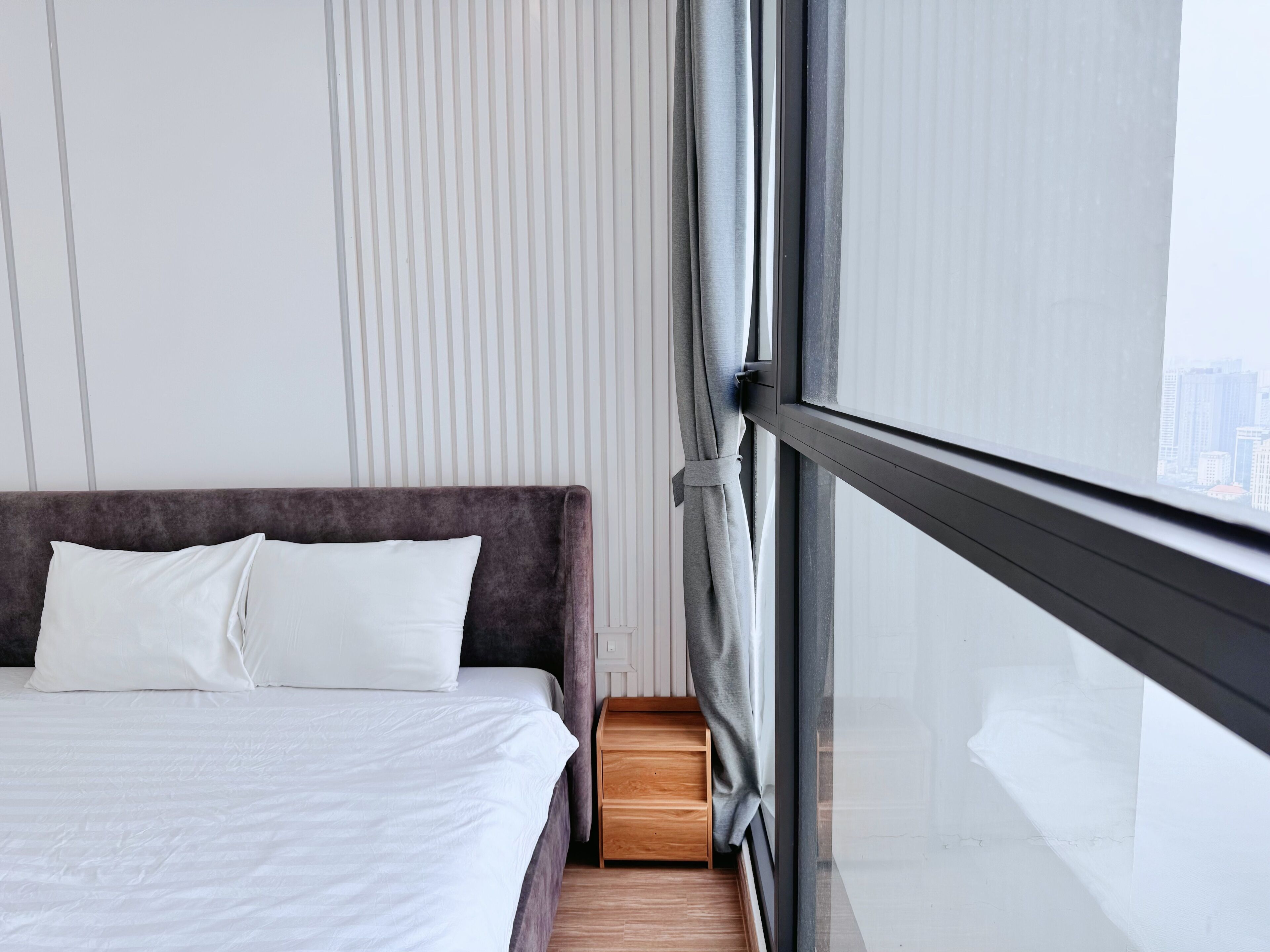 A modern hotel room with a large bed, a wooden nightstand, and a view of the city through floor-to-ceiling windows.