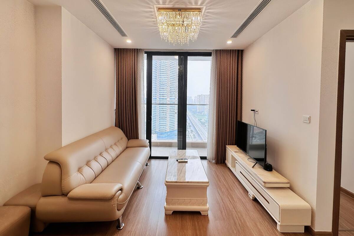 Un salon moderne avec un canapé beige, une table basse, un téléviseur posé sur un meuble en bois et une grande fenêtre avec des rideaux.