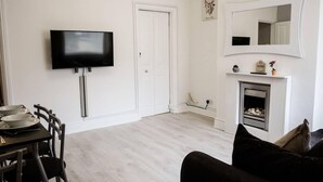 A living room with a flat-screen TV mounted on the wall, a fireplace, a dining area with a table and chairs, and a large mirror above the fireplace. - 4 Bed House, Sleeps 9, Free Parking (Stoke-on-Trent)