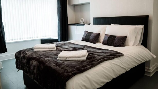 A neatly made bed with a dark brown blanket and white linens, a bedside table with a lamp, and a window with blinds. - 4 Bed House, Sleeps 9, Free Parking (Stoke-on-Trent)