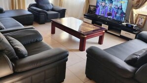 A living room with a large flat-screen TV, black leather sofas, a wooden coffee table, and beige curtains. - 3 Bed House - Sleeps 9 - Pets - Garden - Parking (Accra)