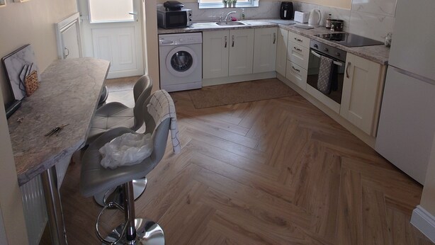 Een kleine keuken met een wasmachine, een spoelbak en een kookplaat. Er is een eethoek met een stoel en een tafel.