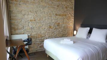A hotel room with a stone wall, a bed with white linens, a desk with a chair, and a nightstand with a lamp.