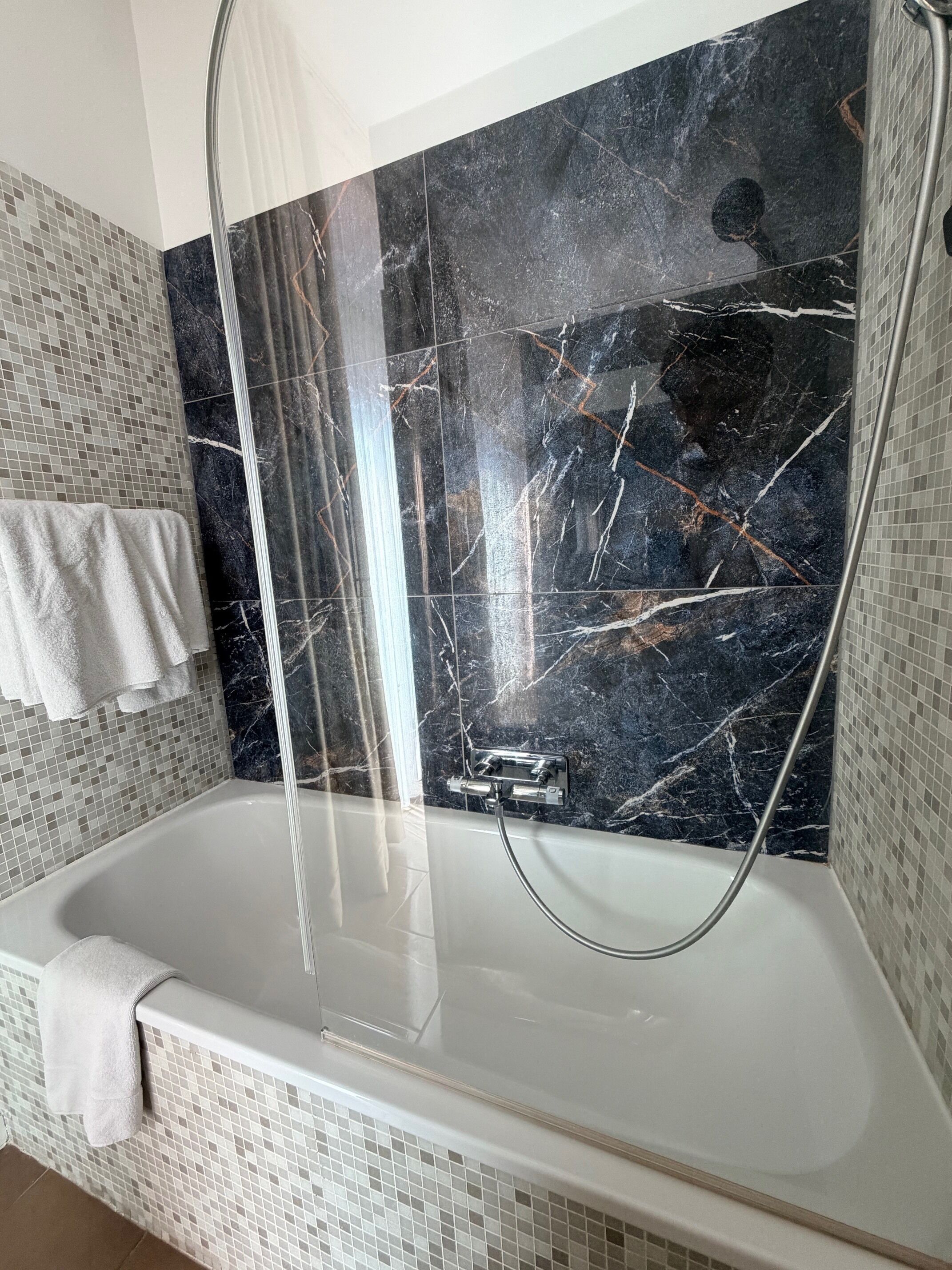A modern bathroom with a marble wall, a glass shower enclosure, and a tiled floor.
