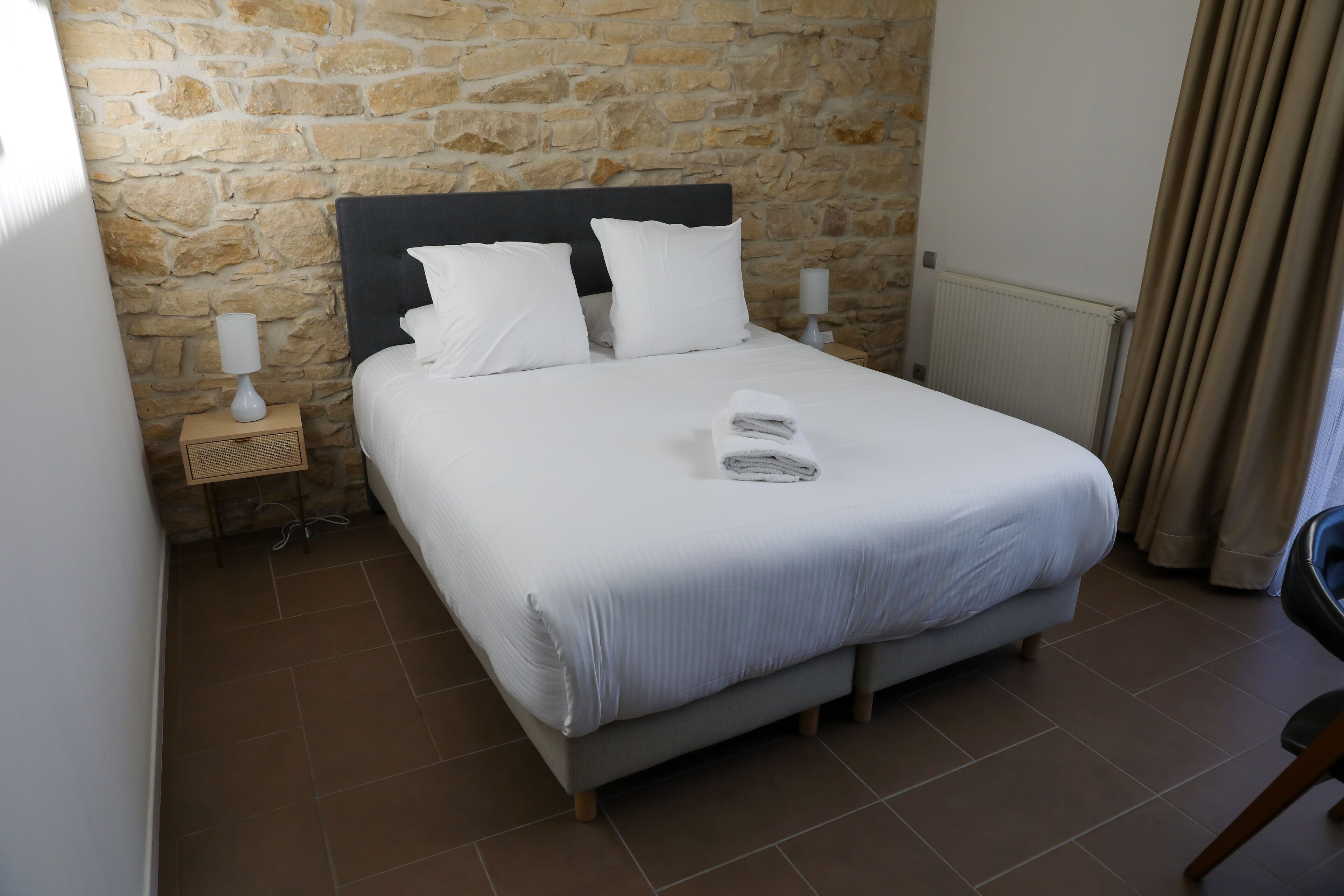 Cama bem arrumada com lençóis e almofadas brancos, mesa de cabeceira com abajur, e parede de pedra ao fundo.