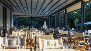 Un'area pranzo all'aperto con tavoli apparecchiati per un pasto, circondata da ampie finestre e alberi.