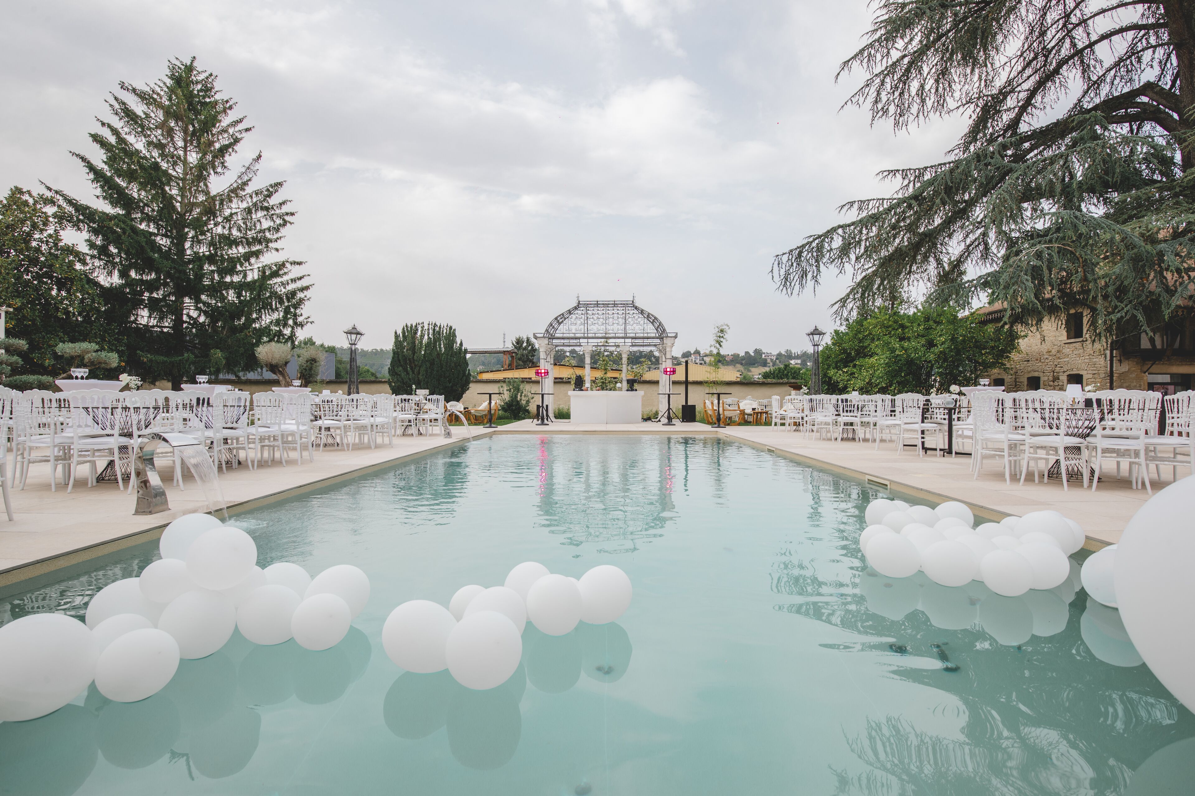 Un área de piscina con sillas y mesas blancas, una pérgola y cielo despejado.