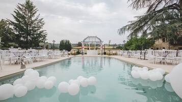 Un área de piscina con sillas y mesas blancas, una pérgola y cielo despejado.