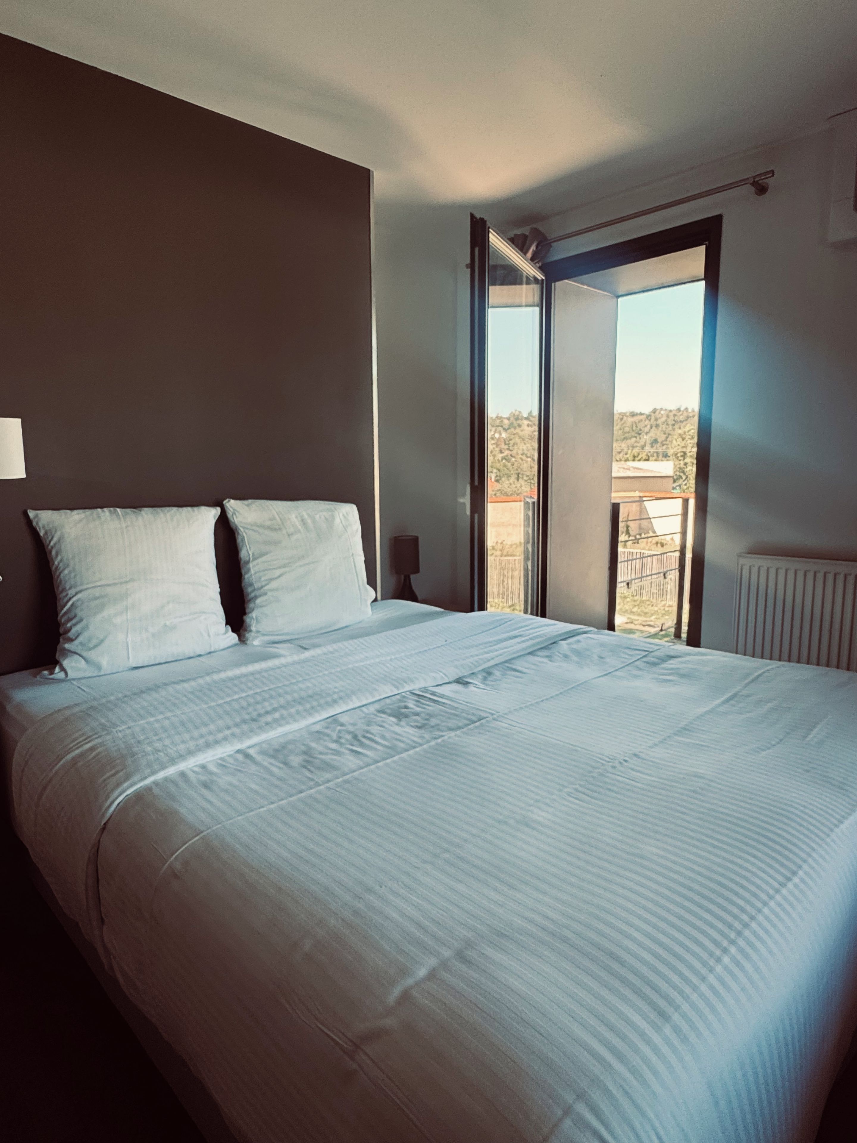 Een hotelkamer met een groot bed, nachtkastje, raam met uitzicht op de omgeving en een radiator.