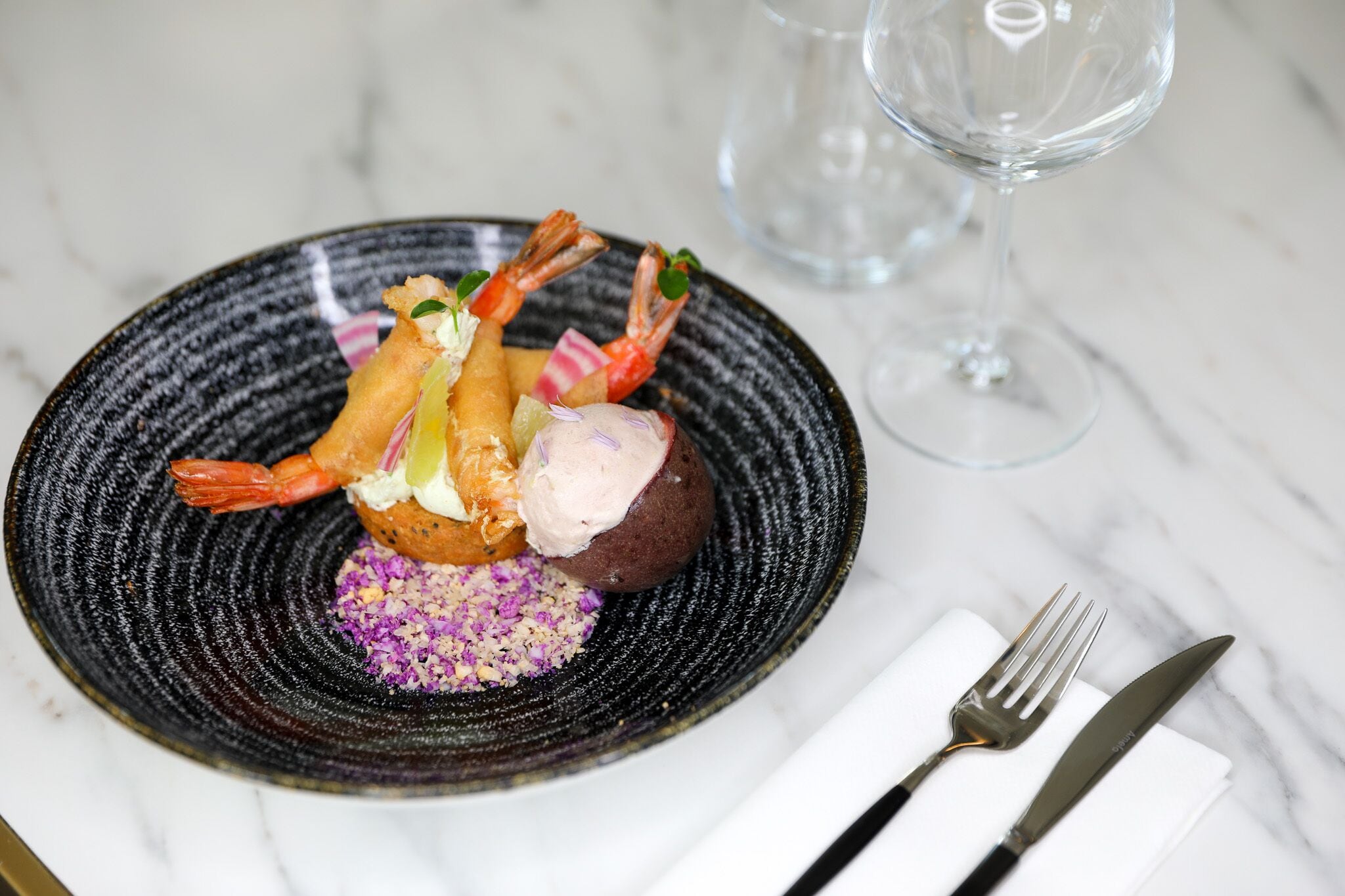 Un plato de postre con camarones, helado y gránulos morados sobre un plato negro texturizado.