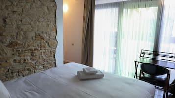 Habitación de hotel con pared de piedra, cama con ropa blanca, escritorio con silla y ventana con cortinas traslúcidas.