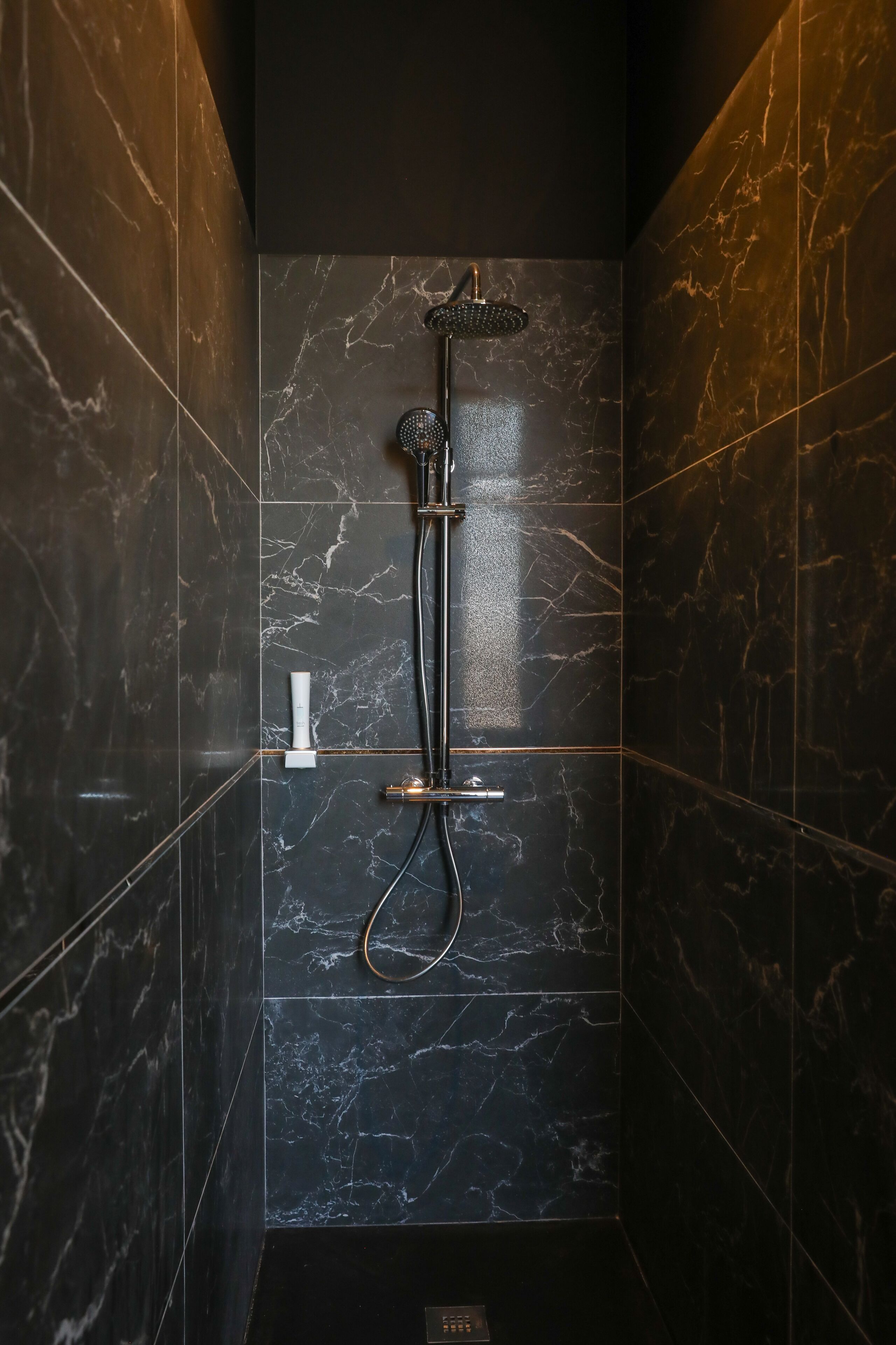 A modern shower with a handheld showerhead and a rain shower head, set against dark marble tiles.