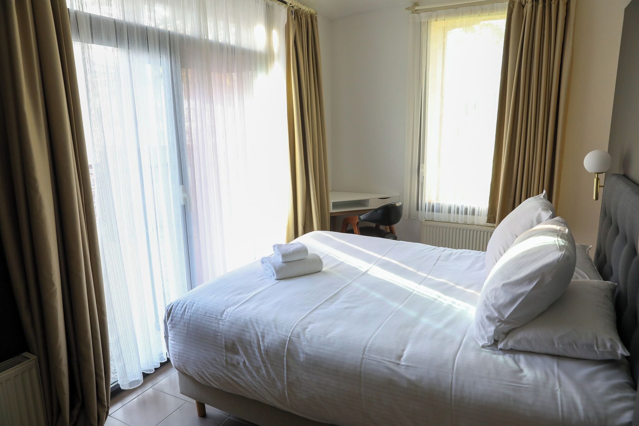 A neatly made bed with white linens and pillows, a bedside table with a lamp, and a window with curtains.