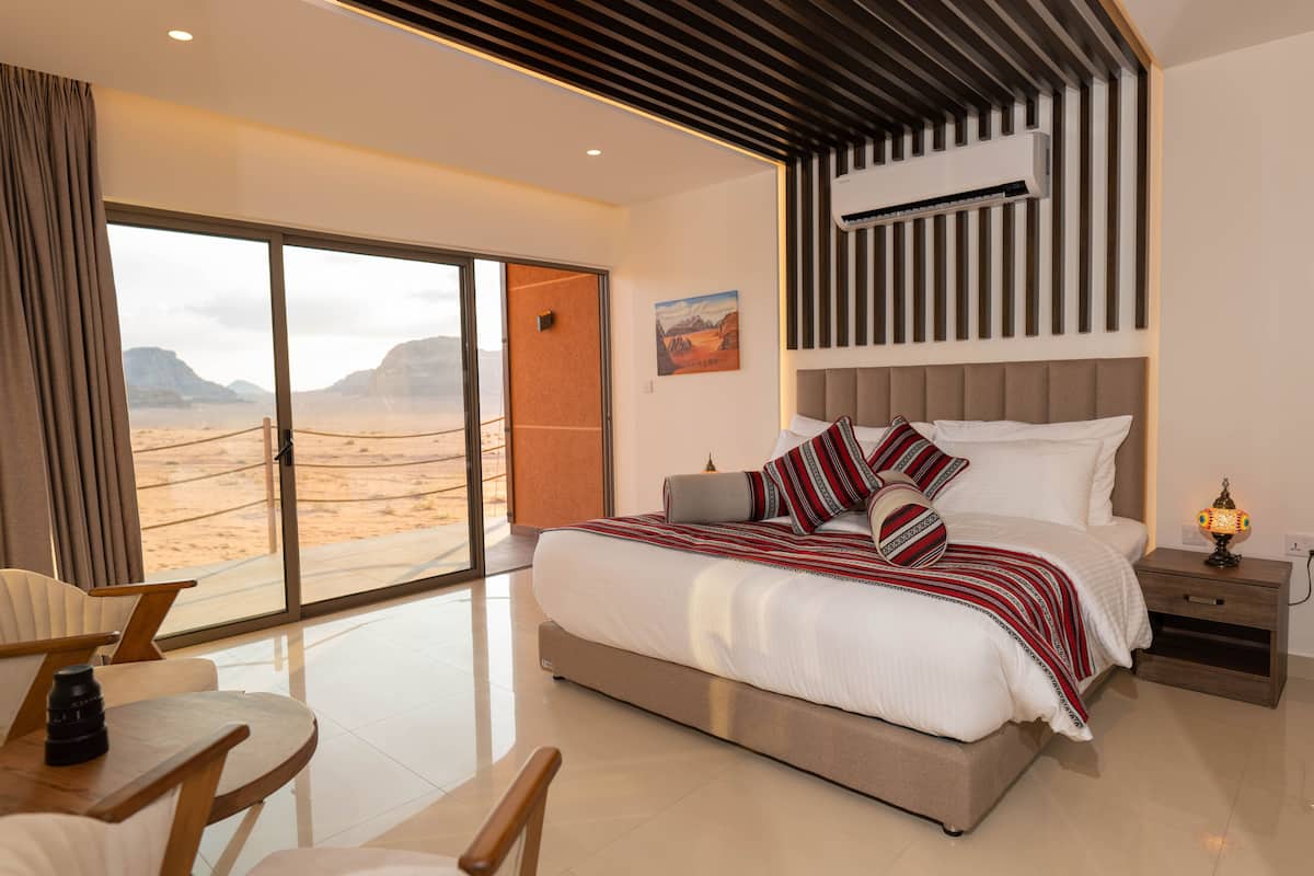 A modern bedroom with a large bed, a nightstand, a lamp, and a view of the desert through a sliding door.