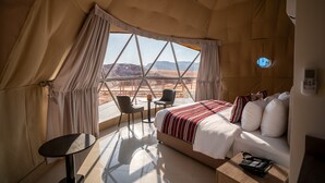 Une pièce aménagée sous une tente, avec un lit, une petite table, une chaise et une vue sur le paysage désertique.