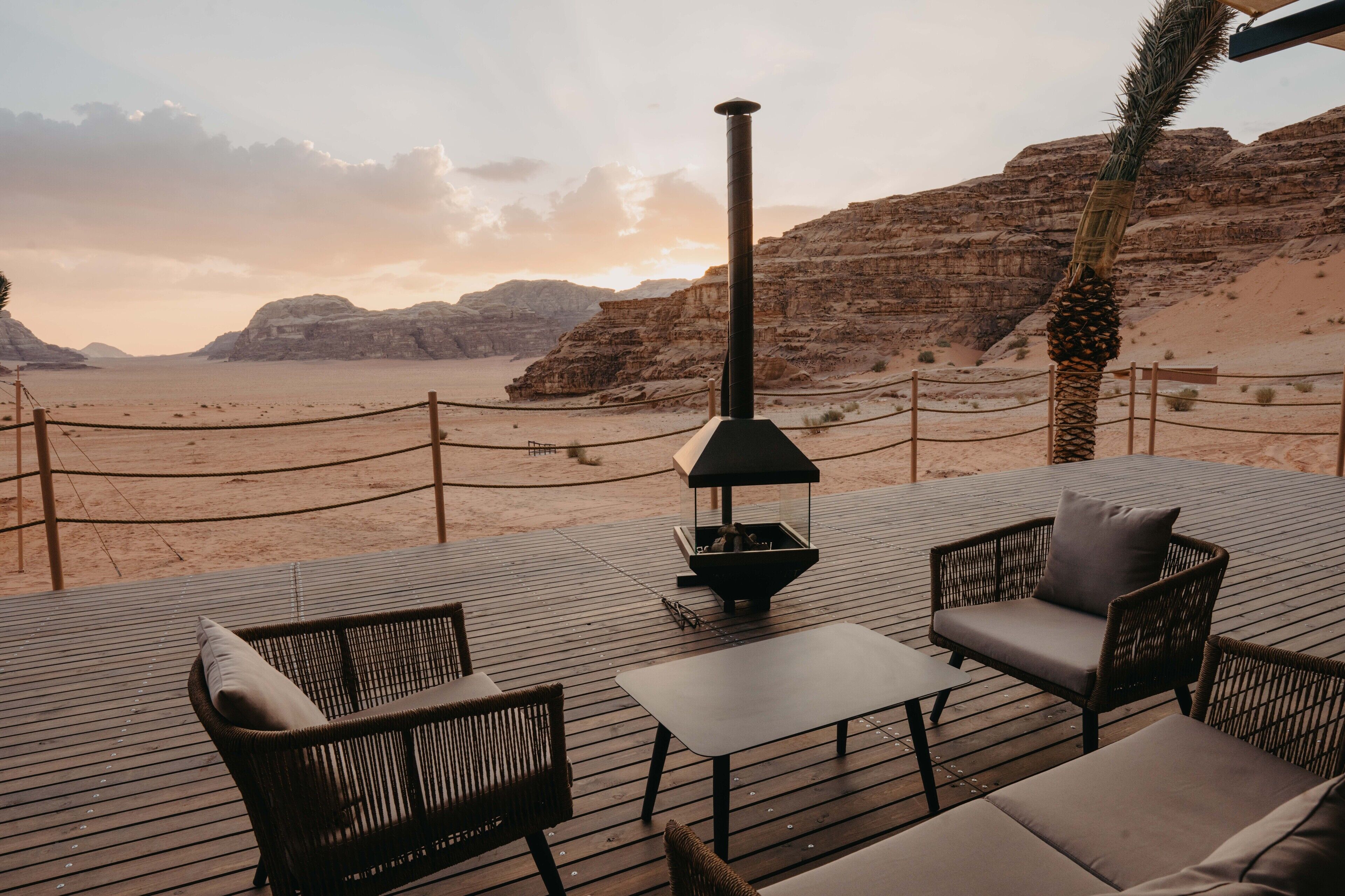 A deck with wicker furniture, a table, and a fire pit, overlooking a desert landscape with rocky hills.
