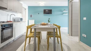 Eine moderne Küche mit weißen Schränken, einer Mikrowelle und einem Backofen. Ein Esstisch mit gelben Stühlen und ein flachbildfernseher an der Wand.