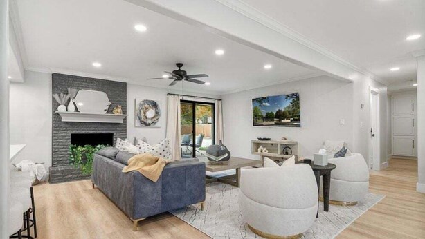 A modern living room with a fireplace, grey sofa, white armchairs, and a ceiling fan.