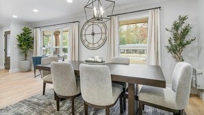 A dining area with a large table, four upholstered chairs, a decorative wall clock, and a hanging light fixture.