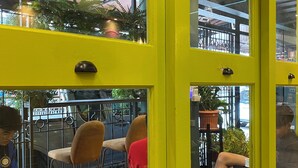 A dining area with yellow walls and a glass door, featuring tables with chairs, a brick floor, and potted plants. - Chomm House (Bangkok)