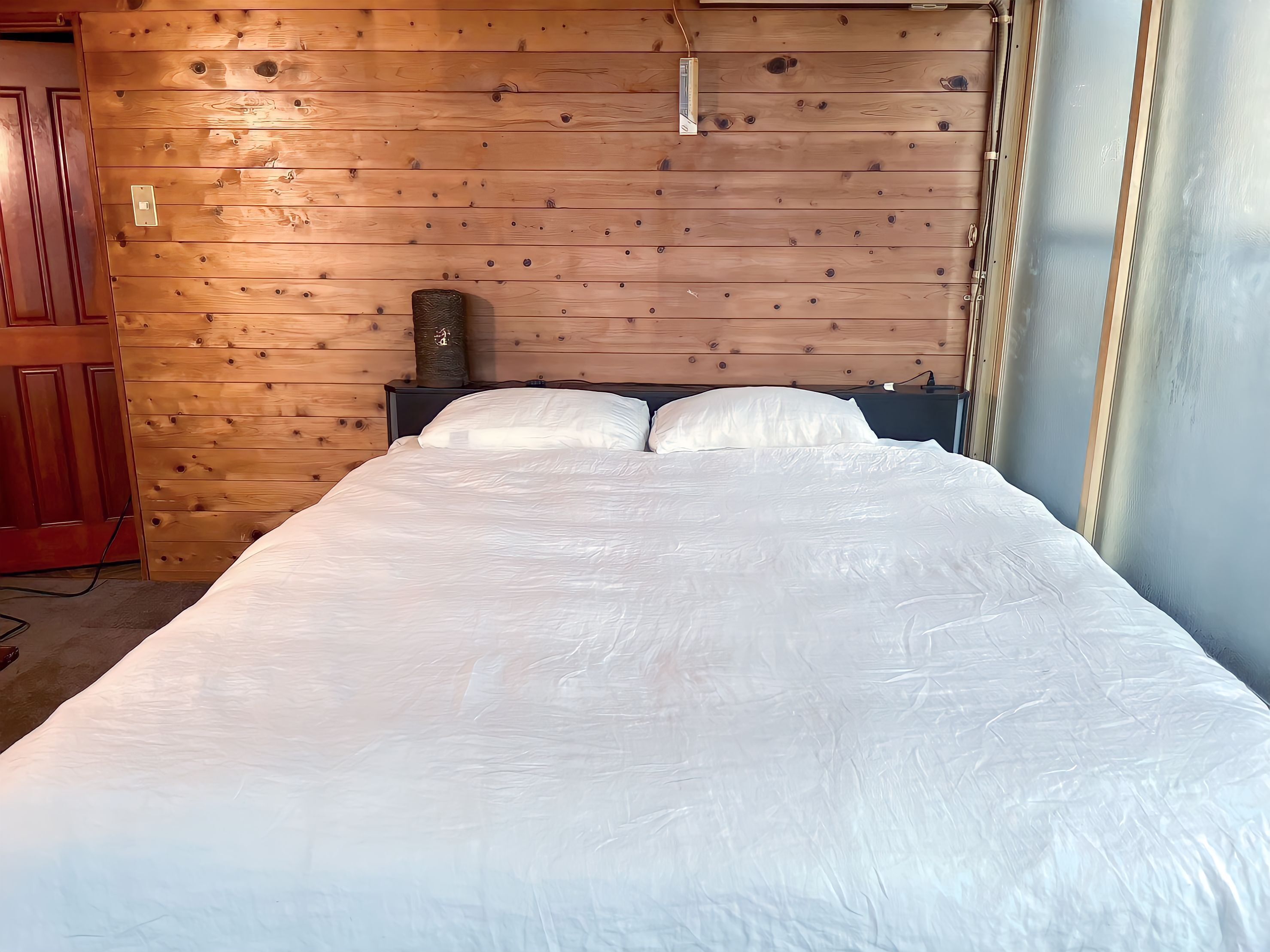 A neatly made bed in a room with wooden panel walls, a visible power outlet, and a door to the left.