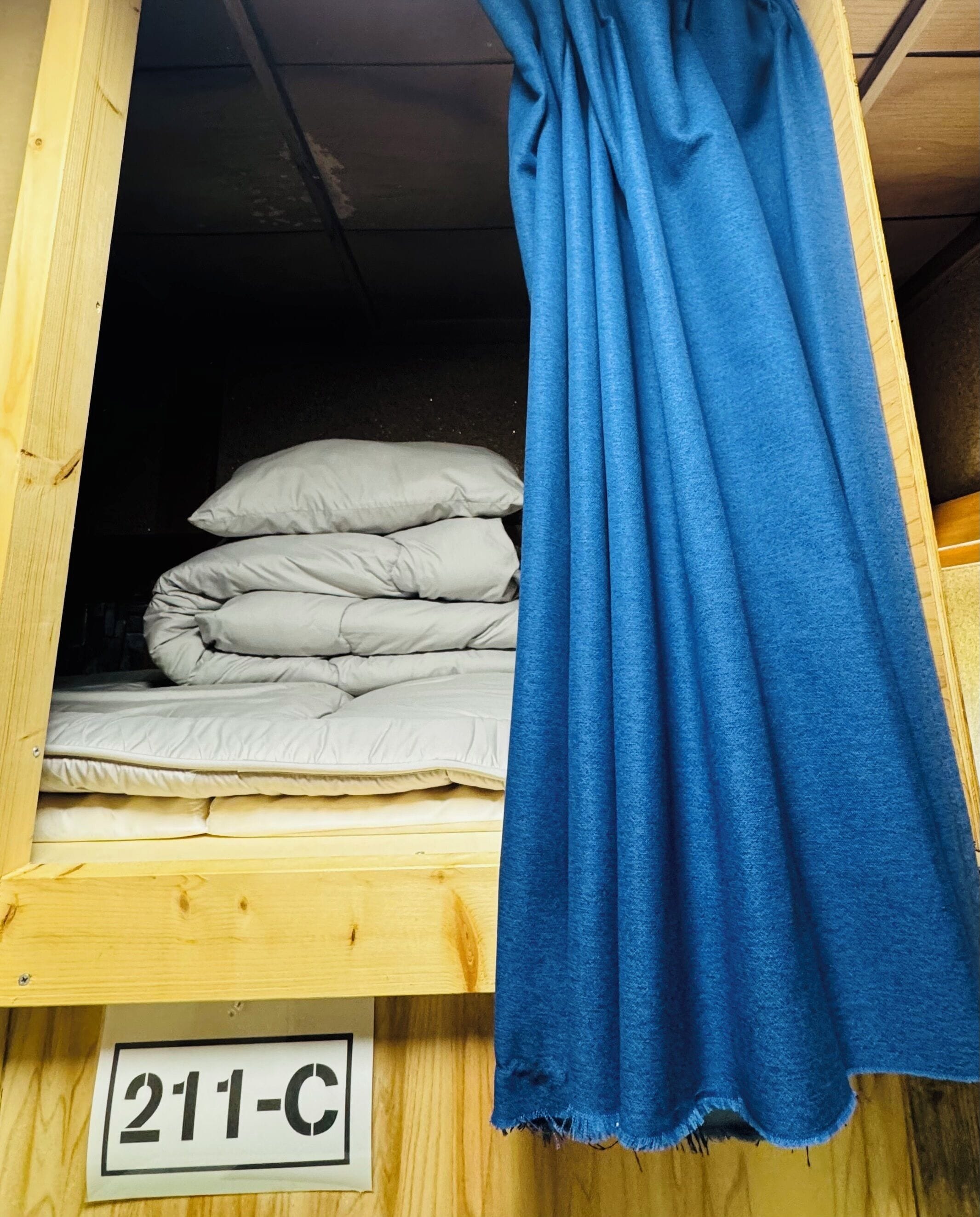 Estructura de cama de madera con colchón y almohadas, una cortina azul y un cartel con el número 211-C.