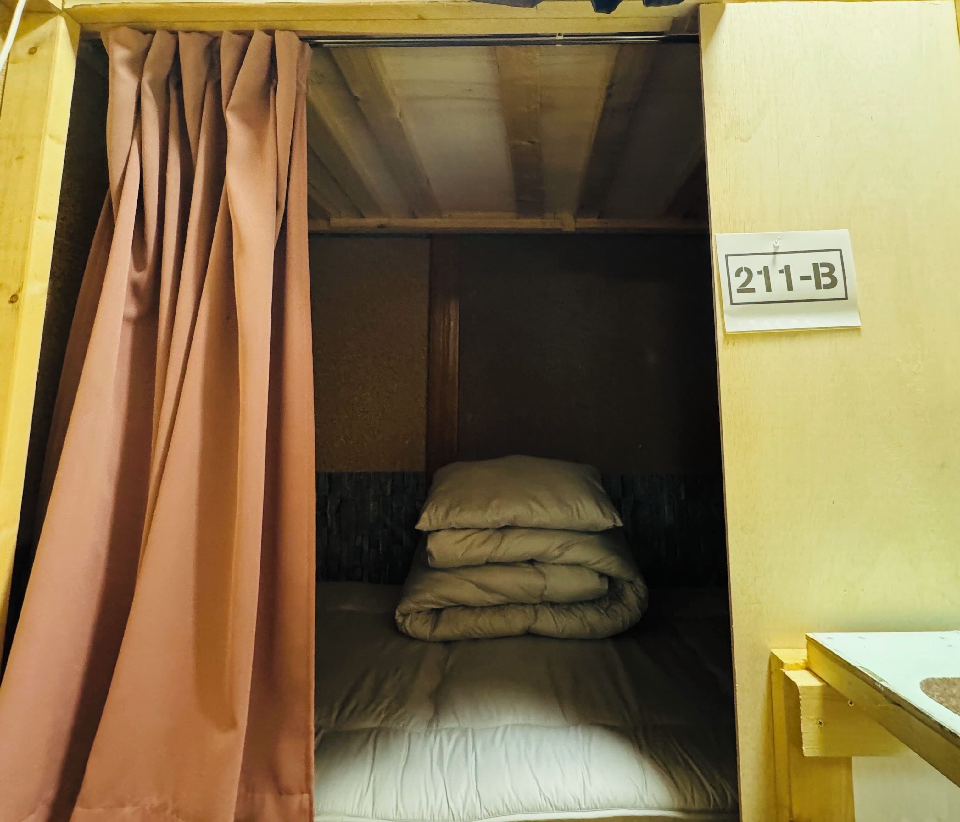 Un lit simple avec une couverture pliée, un oreiller et une pancarte indiquant le numéro de chambre : 211-B.
