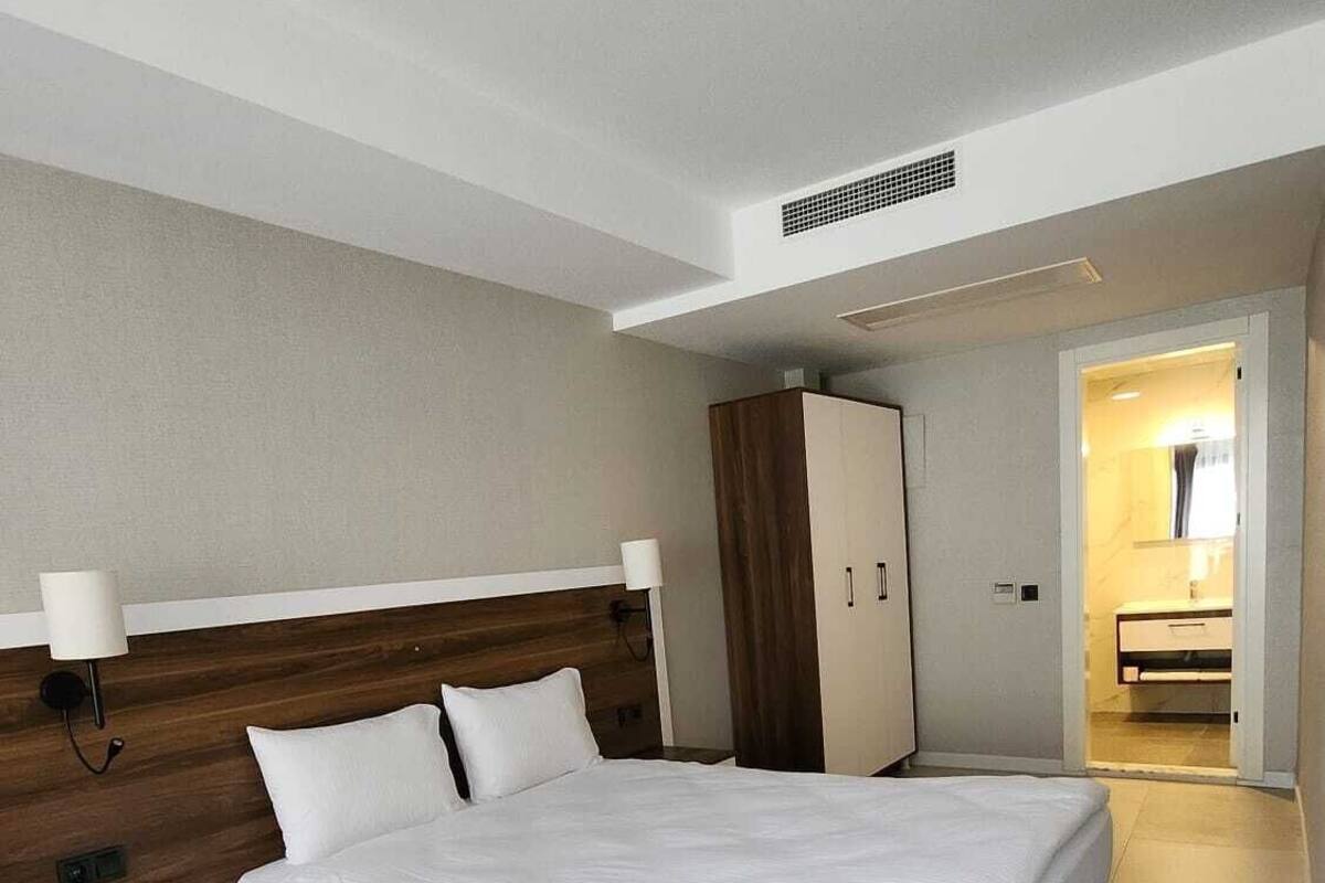 A modern hotel room with a large bed, a wooden headboard, a nightstand, a wardrobe, and a bathroom visible through an open door.