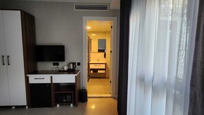 Habitación de hotel compacta con kitchenette, televisor montado en la pared, escritorio pequeño con cafetera y baño visible a través de una puerta abierta.