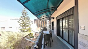 Un balcón con mesa y sillas, un perro, y vistas de edificios y árboles cercanos.