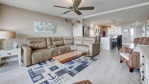 A modern living room with a beige sectional sofa, a wooden coffee table, and a patterned rug. There is a ceiling fan, a dining area with chairs, and a kitchen in the background. - 404 Ocean View Condo (Avalon Beach Club Assn) (Fort Pierce)