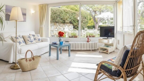 Ein helles Wohnzimmer mit einem weißen Sofa, einem Korbstuhl, einem kleinen Tisch mit einer Vase und einem Fernseher auf einem Lowboard.