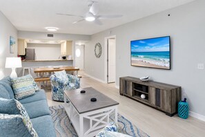A living room with a sofa, coffee table, and TV. There is a kitchen in the background.