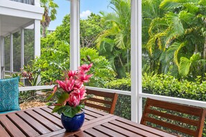 A wooden table with a blue pot of pink flowers, a blue patterned pillow, and a view of lush greenery through large windows. - Garden view Siesta Key condo Pickleball, 2 pools, beach access - GBB 1326 (Siesta Key)