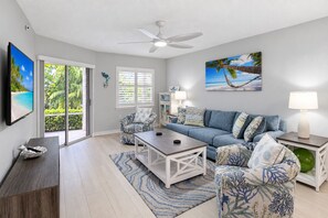 A spacious living room with a large sofa, a coffee table, and a television. There is a sliding glass door leading to a balcony with a view of palm trees.