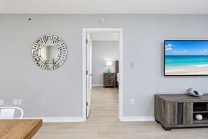 A room with a wooden dining table, a TV displaying a beach scene, a mirror with a geometric design, and a hallway leading to another room. - Garden view Siesta Key condo Pickleball, 2 pools, beach access - GBB 1326 (Siesta Key)