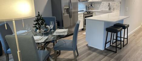 Cocina moderna con armarios blancos, refrigerador de acero inoxidable, microondas y un lavamanos. Hay una mesa de comedor con sillas y un pequeño árbol de Navidad.