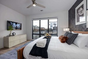 Ein modernes Hotelzimmer mit einem großen Bett, einem Deckenventilator, einem Fernseher, einem Fenster mit Blick auf Gebäude und einer Pflanze auf dem Nachttisch.