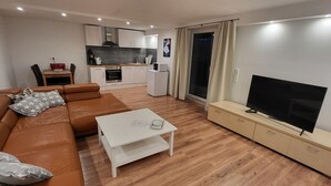 Un salon moderne avec un canapé en cuir marron, une table basse blanche, un téléviseur posé sur un meuble en bois, et une cuisine en arrière-plan.