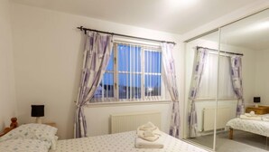 A bedroom with a large bed, a window with curtains, a radiator, a mirror, and a bedside table with a lamp. - Cedar House by Interhome (Inverness)