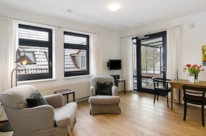 Una habitación con sofá, sillón, reposapiés, una mesa con sillas, televisor y ventanas amplias con cortinas.