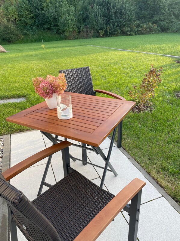 Une terrasse extérieure avec une table et des chaises en bois, entourée d’une pelouse verdoyante et d’arbres.