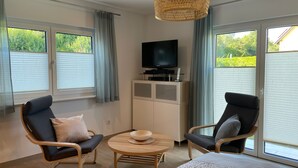 Ein modernes Zimmer mit zwei Sessel, einem kleinen Tisch, einem Fernseher auf einer Kommode und großen Fenstern mit Jalousien.