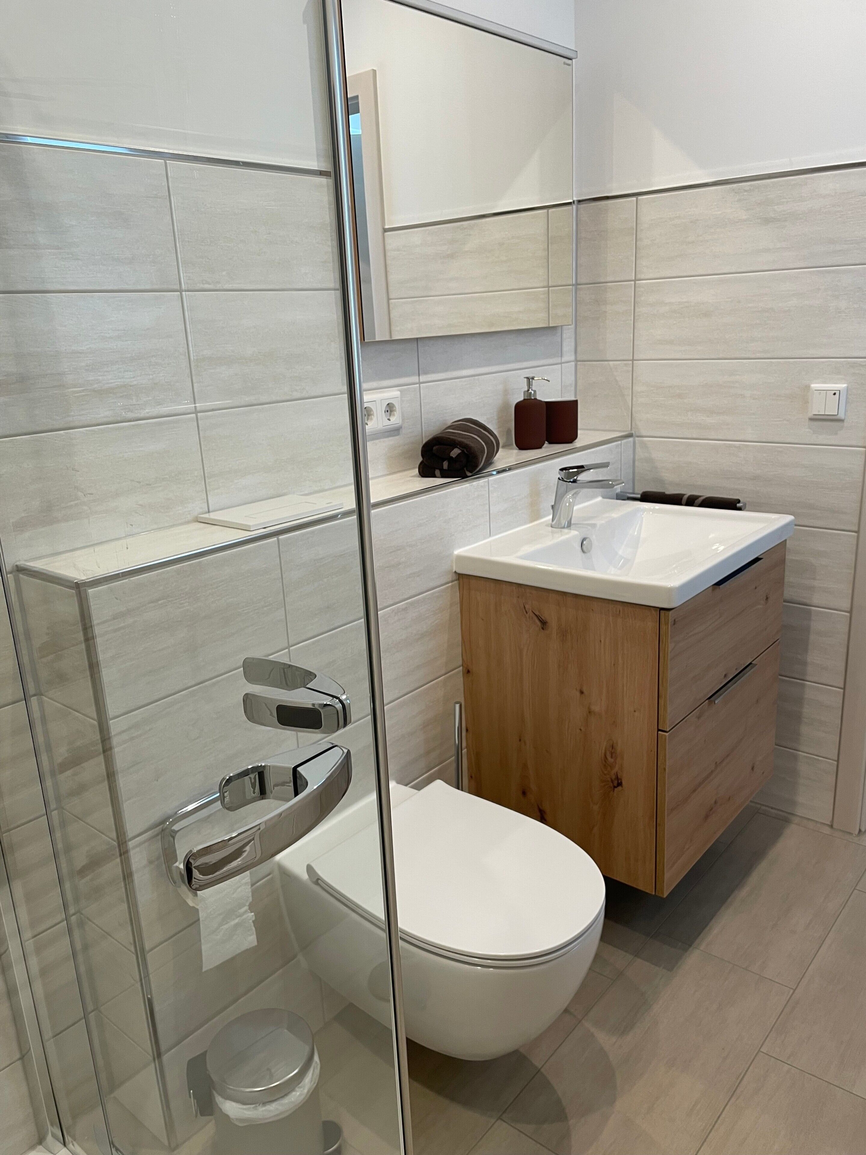 Et moderne badeværelse med et glasbadeværelsesafskærmning, et hvidt toilet, et trækommode med vask og et spejl over kommoden.